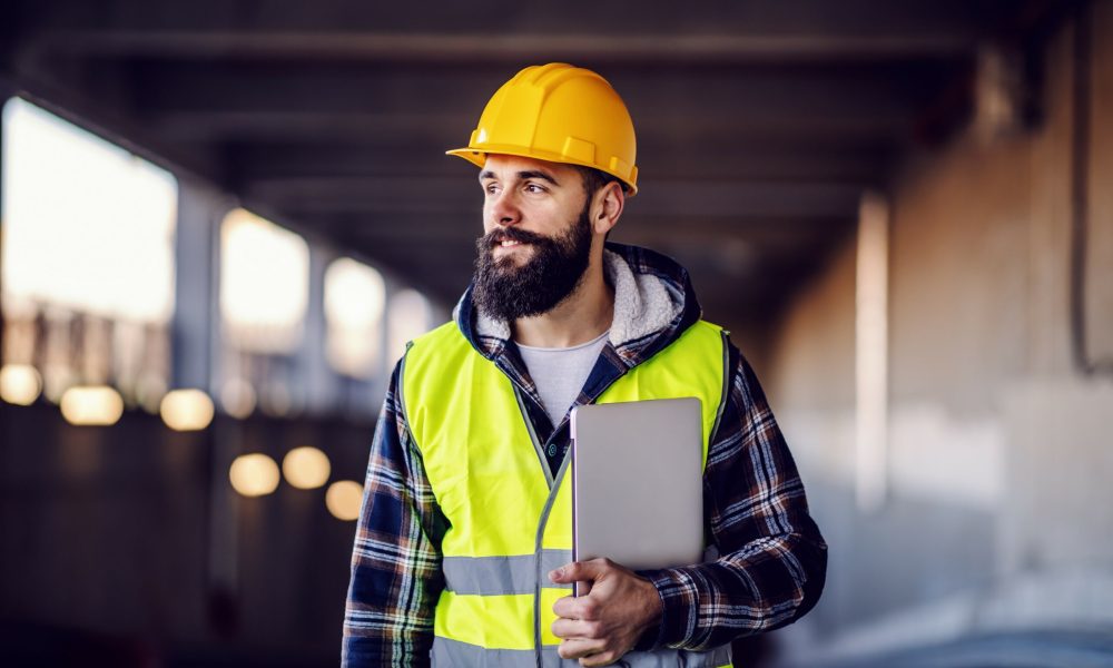 A construction worker with a beard, wearing a yellow hard hat and high-visibility vest, holds a tablet and looks off into the distance.