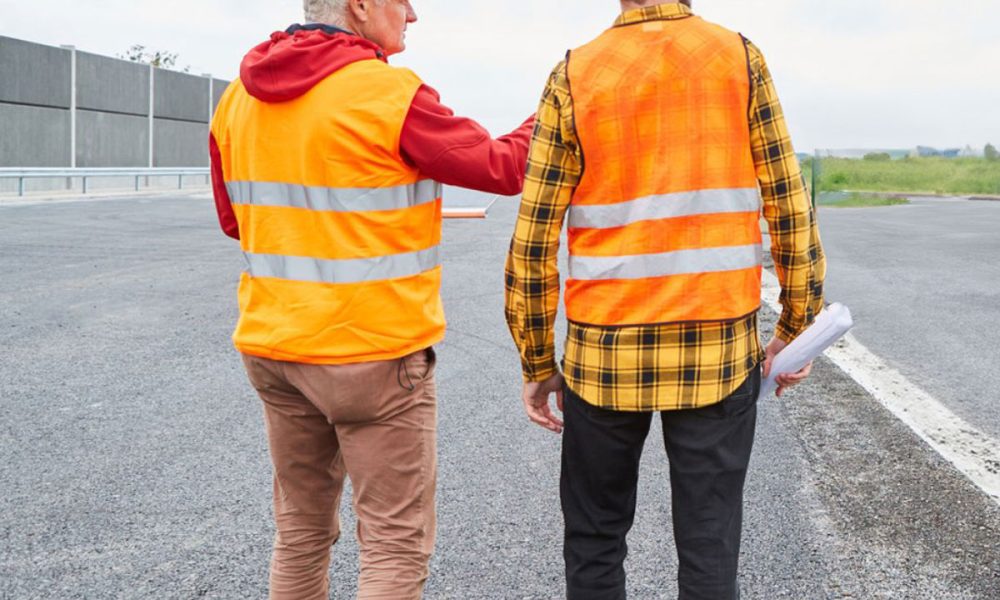 Two construction workers in high-visibility vests and hard hats discussing on a newly paved road.